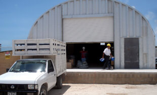 Un almacén metálica con una puerta de garaje partialmente abierta y un carro delante, un hombre con casco amarilla camina delante