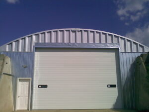 Una vista frontal de una estructura arqueada de acero. Es el Modelo S de SteelMaster. La pared de extremo tiene una puerta de garaje muy grande y una puerta de servicio al lado de la.