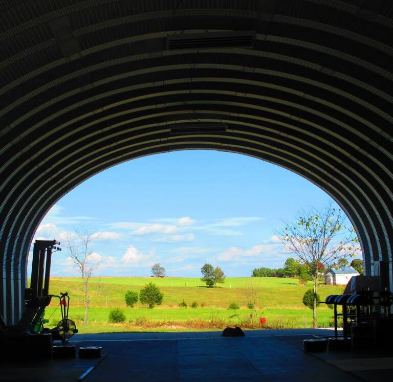 Una vista interior de un gimnasio de crossfit dentro de una estructura arqueada de acero. No hay una pared de extremo y puedes ver una vista hermosa de la tierra verde y cielo azul.
