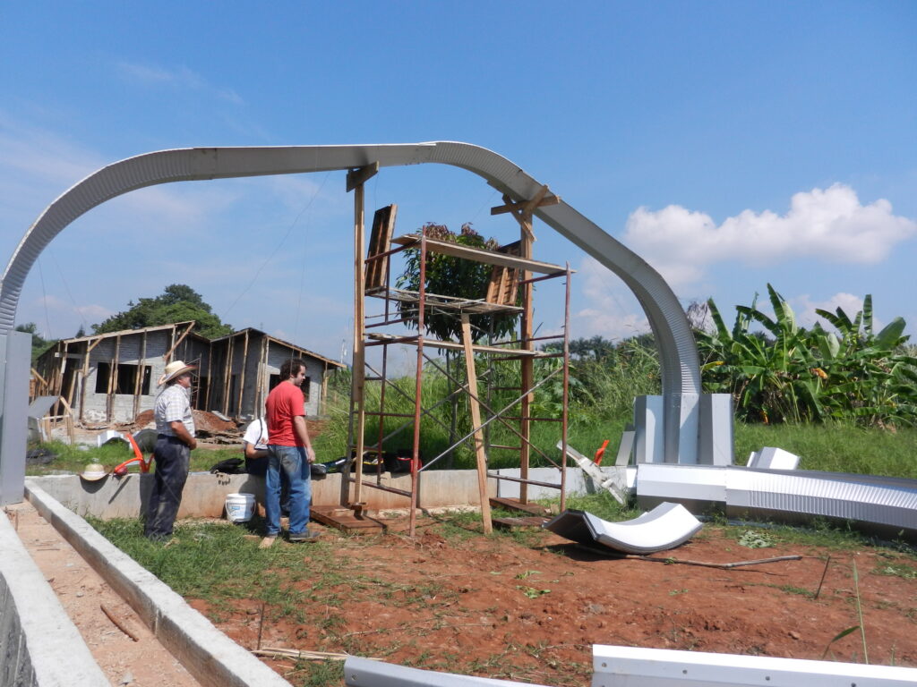 La construcción de una estructura del Modelo A SteelMaster. Hay personas en ropa normal que están armando los arcos de acero. Hay piezas de arco en el piso.