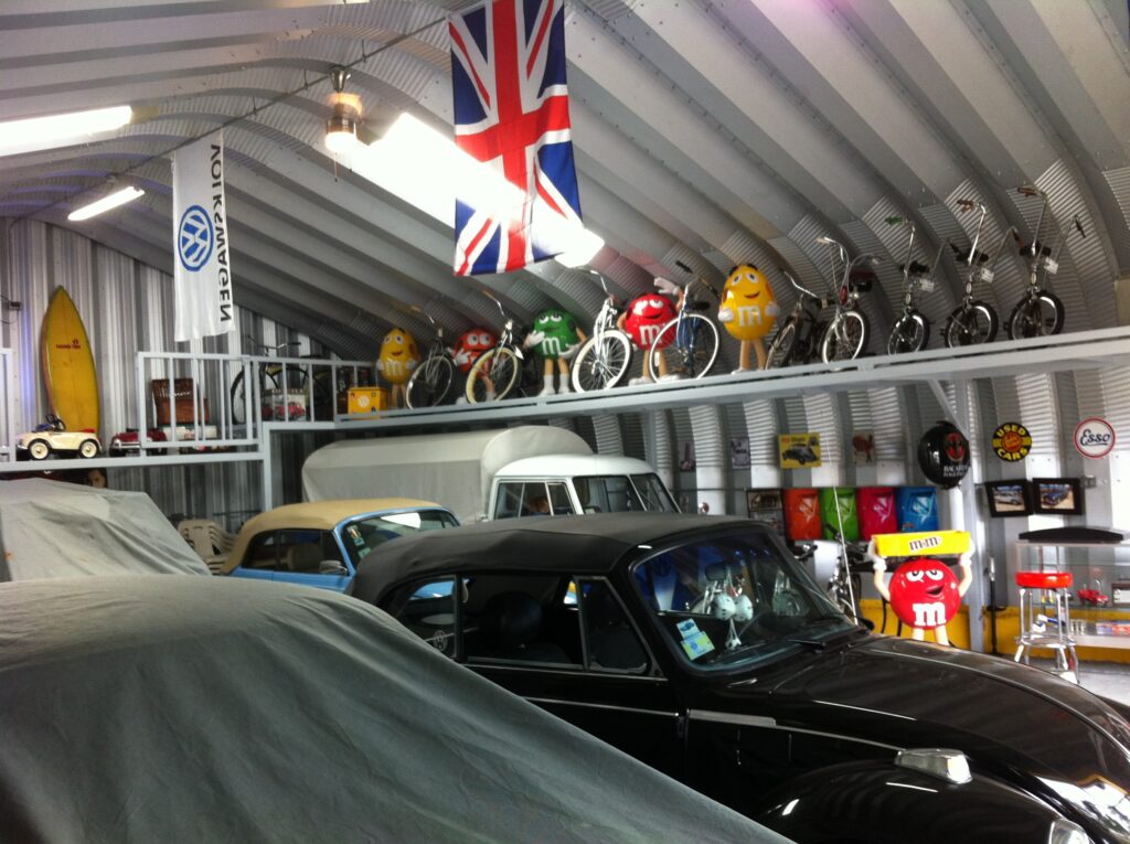 Una vista interior de un garaje con coches antiguos y otras antigüedades. Hay un coche negro y una bandera de Inglaterra. En los estantes de la pared hay una colección de bicicletas miniatura y figuras de M&M.