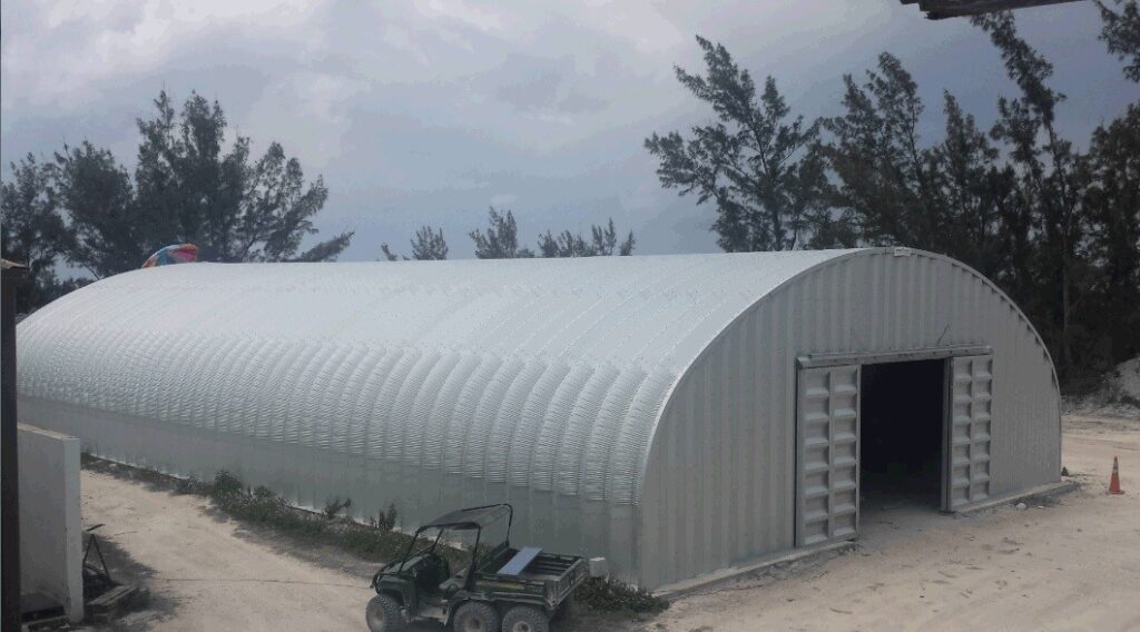 Un almacén de acero. La estructura es arqueada con una puerta grande en la pared de extremo. En el fondo hay arbóles tropicales.