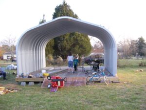 Una estructura arqueada de acero del Modelo A SteelMaster en construcción. Los arcos están armados; no hay paredes de extremo. Hay herramientas en el piso y hay dos personas debajo de la estructura.