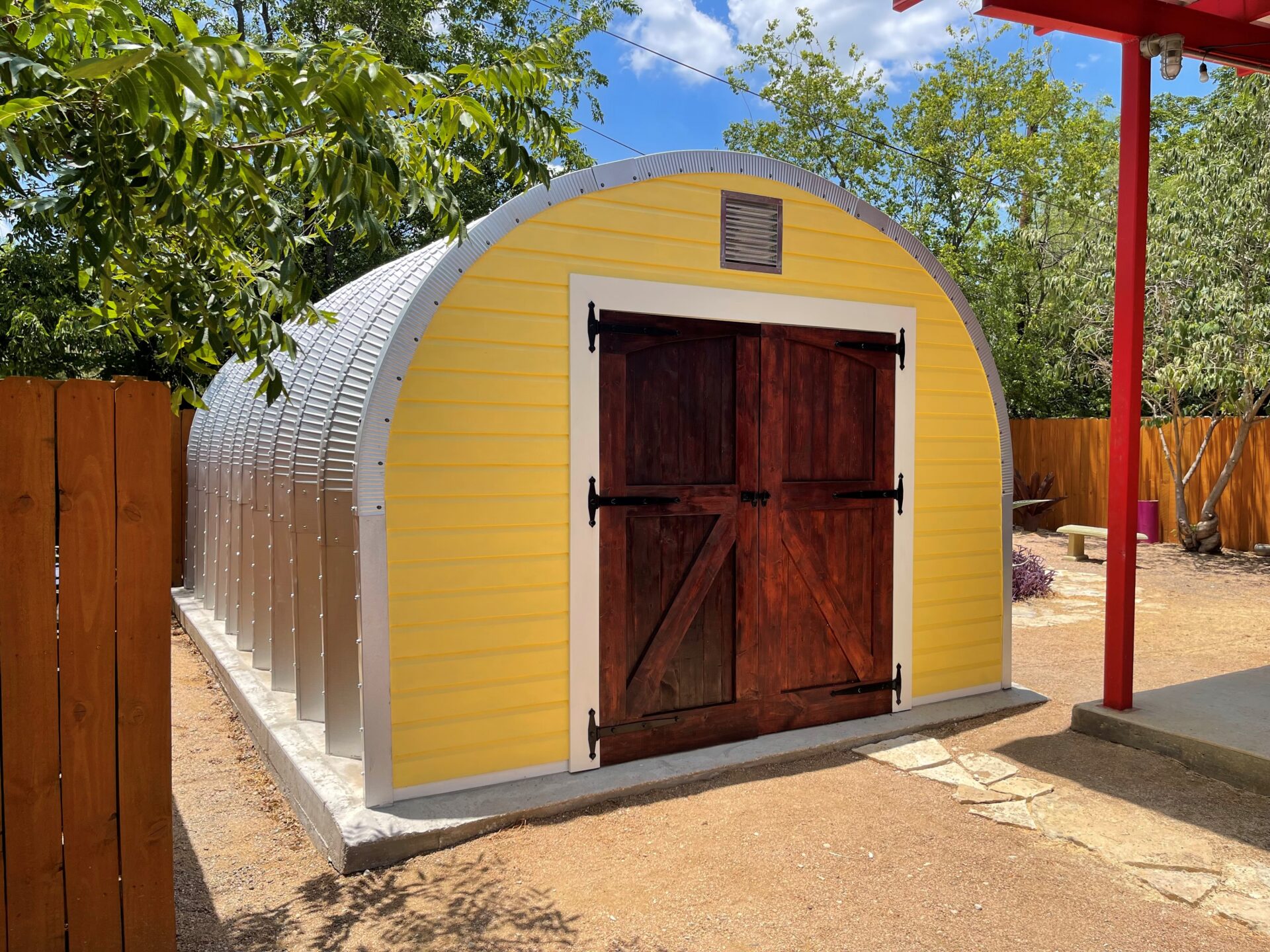 Un cobertizo residencial de Modelo S. La pared de extremo es personalizada y tiene una puerta de madera y está pintada de amarillo. En el fondo hay una valla y árboles.