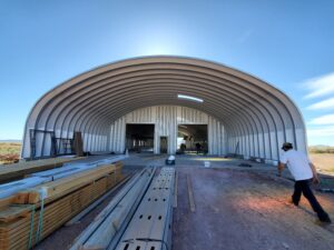 Una estructura arqueada de acerp. Está en construcción. Hay materiales de construcción en el primer plano y un hombre está andando a la estructura.