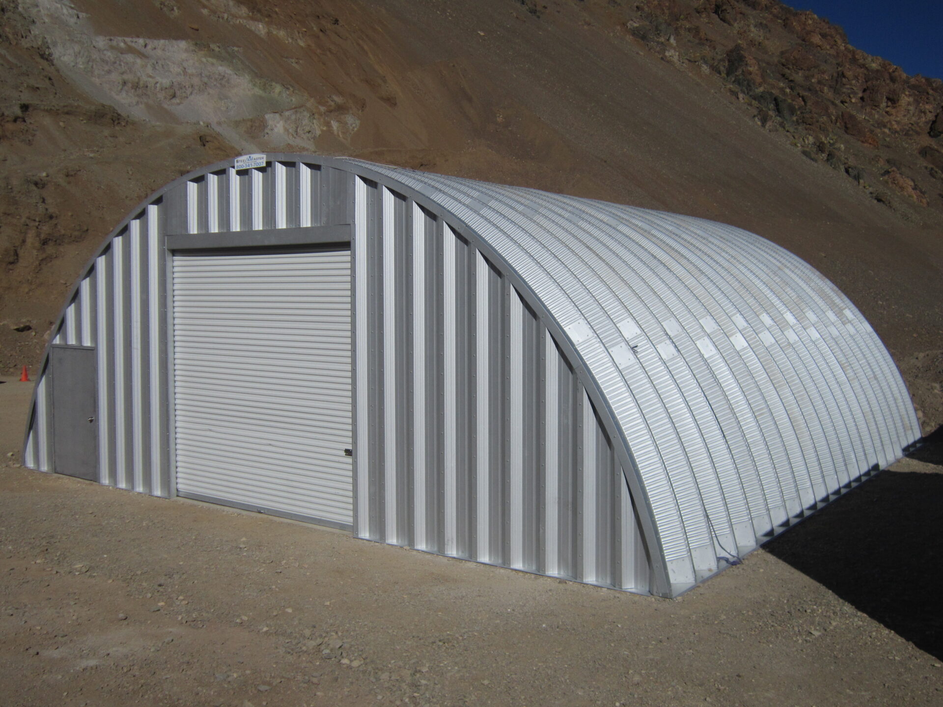 En el primer plano hay una estructura de acero. Es el modelo Q de SteelMaster. Este Quonset Hut tiene dos puertas. Una es grande para vehículos y la otra es más pequeña para personas. En el fondo, hay montañas y una escena bonita del desierto.