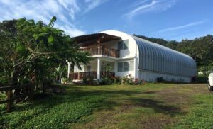Una casa blanca de estilo arco. Usa el modelo S de SteelMaster y tiene una pared de extremo personalizada. Tiene dos pisos y una terraza en el segundo piso que está encima de un porche en el primer piso. Hay un jardín con muchas plantas, flores y árboles. También en la pared de extremo hay ventanas.