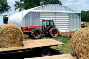 En el primer plano hay un tractor y dos balas de heno. En el fondo hay una estructura de acero que tiene algún uso de agrícola. La estructura es el Modelo S de SteelMaster.