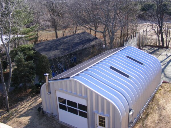 Una foto de un garaje o cochera que tiene paneles solares en el techo. La estructura es de acero y tiene una puerta de cochera y una de servicio. También hay una chimenea pequeña y dos tragaluces. El garaje es sostenible y bueno para el medio ambiente porque usa energía solar. Está ubicado en una escena rural con árboles.