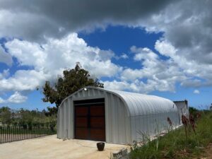 Una estructura arqueada de acero. Es el modelo A SteelMaster. En la pared de extremo, hay una puerta de garaje que imita la madera.