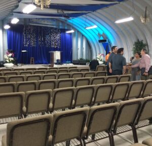 Vista interior de una iglesia con techo arqueado de acero. El espacio cuenta con decoraciones azules, filas de sillas marrones en primer plano y personas conversando en el pasillo